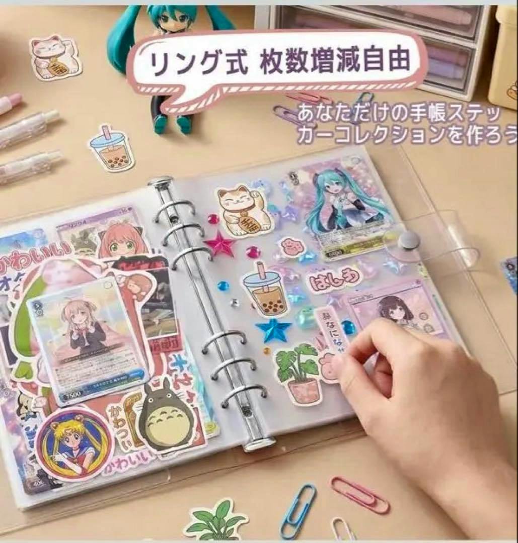 An open clear ring binder with A4 plastic sleeves holding stickers and cards on a desk, with a hand arranging a sticker.
