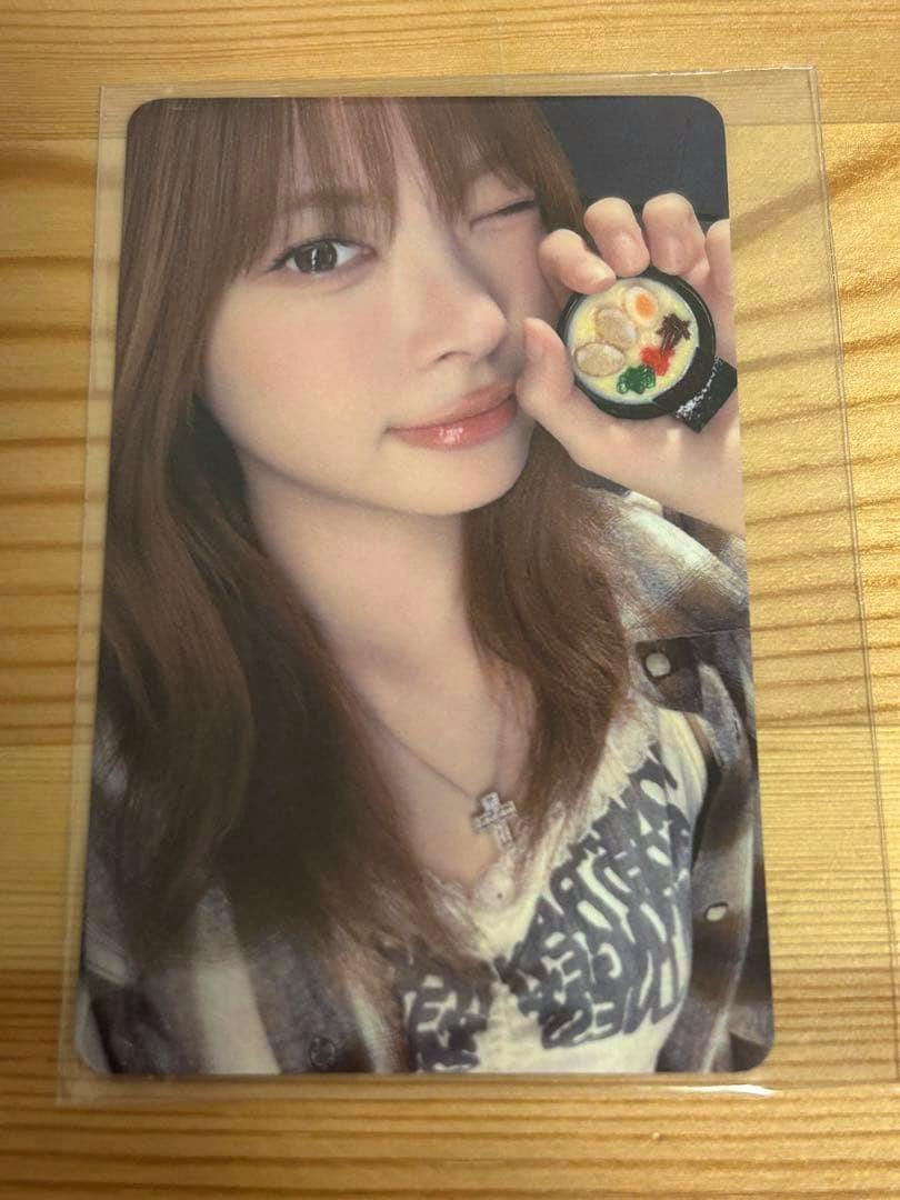 Photocard showing a close-up of a woman winking and holding a small bowl prop, inside a plastic sleeve on a wooden surface.