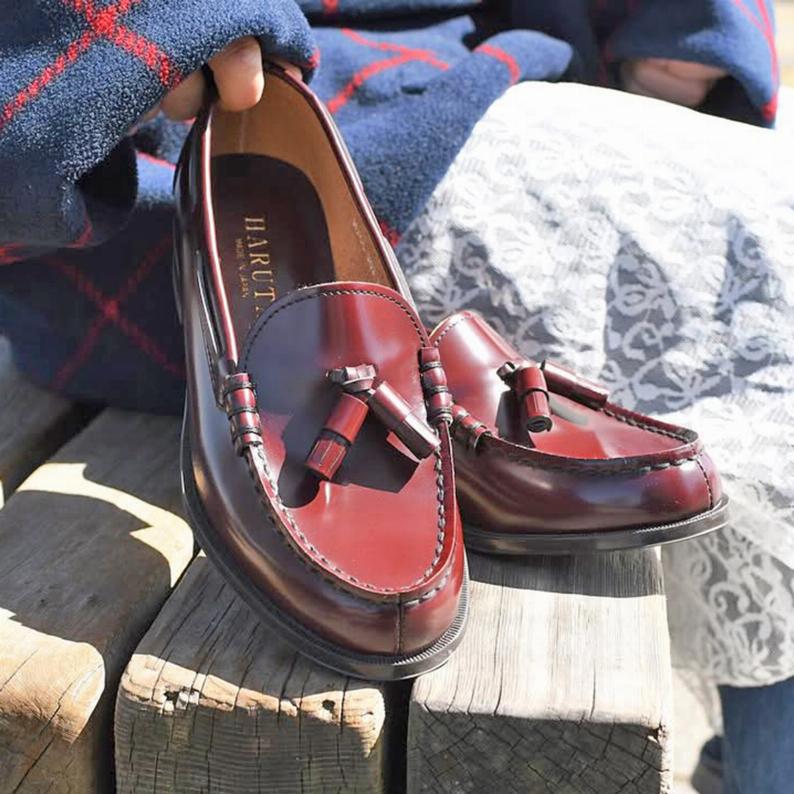Close-up of burgundy leather tassel loafers on a wooden bench, with one shoe held by a person.
