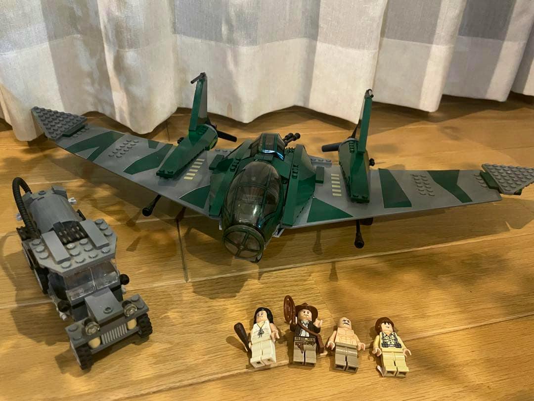 Assembled LEGO green aircraft, a gray vehicle, and four minifigures from the Indiana Jones set displayed on a wooden floor.