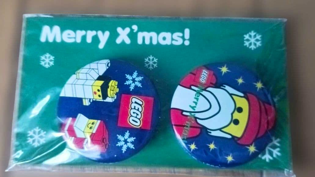 Packaged LEGO Christmas two-can-badge set on green backing showing two round badges with LEGO minifigure faces and the text "Merry X'mas!".