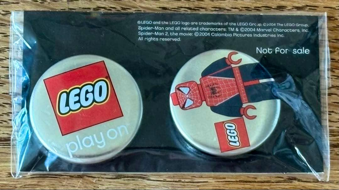 Two round metal LEGO badges in sealed plastic packaging on a black backing, one showing the LEGO logo and the other showing a Spider-Man minifigure graphic.