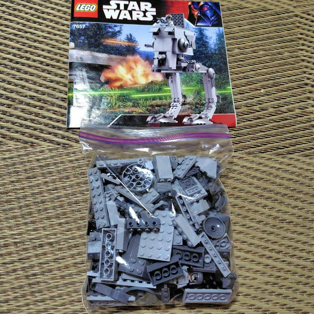 Resealable plastic bag of light gray LEGO pieces shown below the LEGO Star Wars 7657 AT-ST instruction sheet on a woven surface.