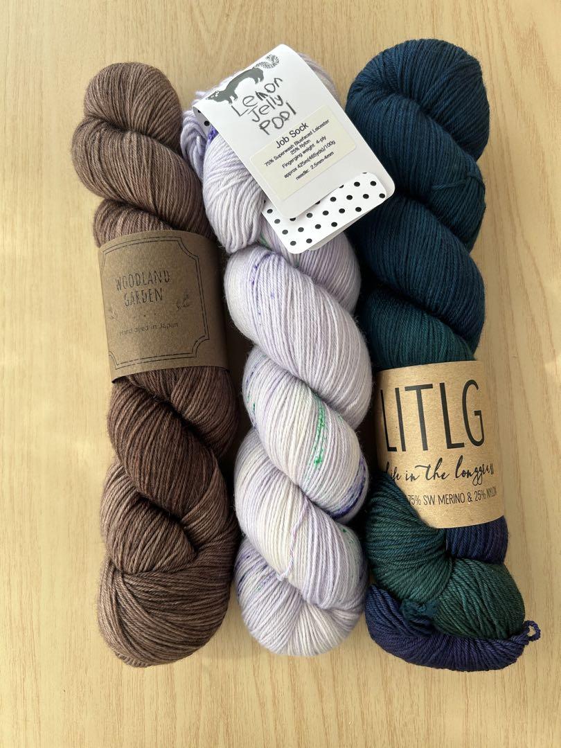 Three hand-dyed skeins of sewing thread (cotton) in brown, light purple, and dark teal laid side-by-side on a wooden surface.
