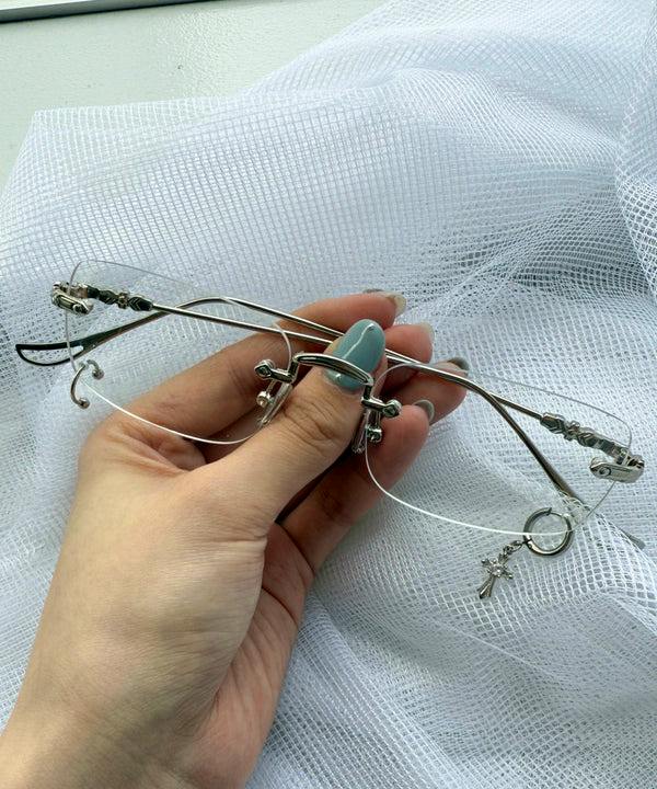 A hand holds rimless eyeglasses with silver metal temples and small hanging charms against a white mesh background.
