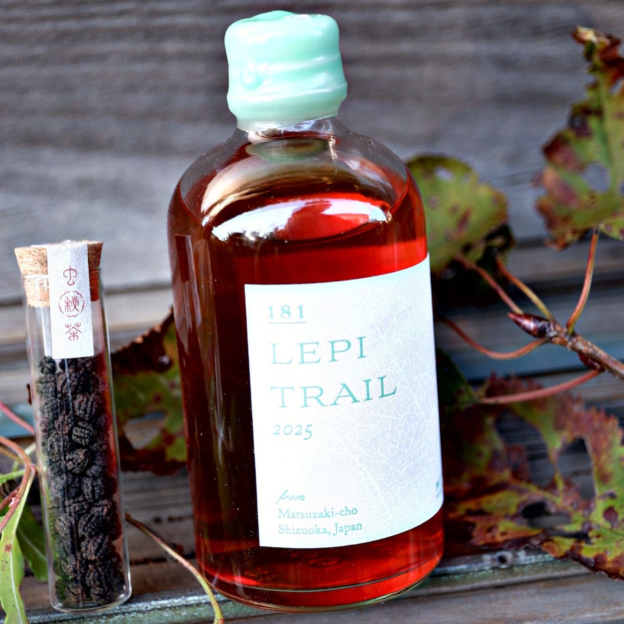 A glass bottle labeled "LEPI TRAIL" alongside a small glass vial of loose tea displayed on a wooden surface with leaves.