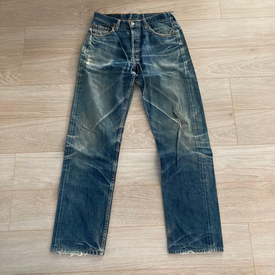 Front view of worn Levi's 501 denim jeans laid flat on a wooden floor, showing whiskering and frayed hems.