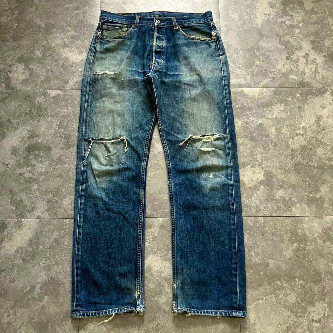 Front view of men's Levi's 501 blue denim jeans laid flat on a tile floor, showing faded wash and knee rips.