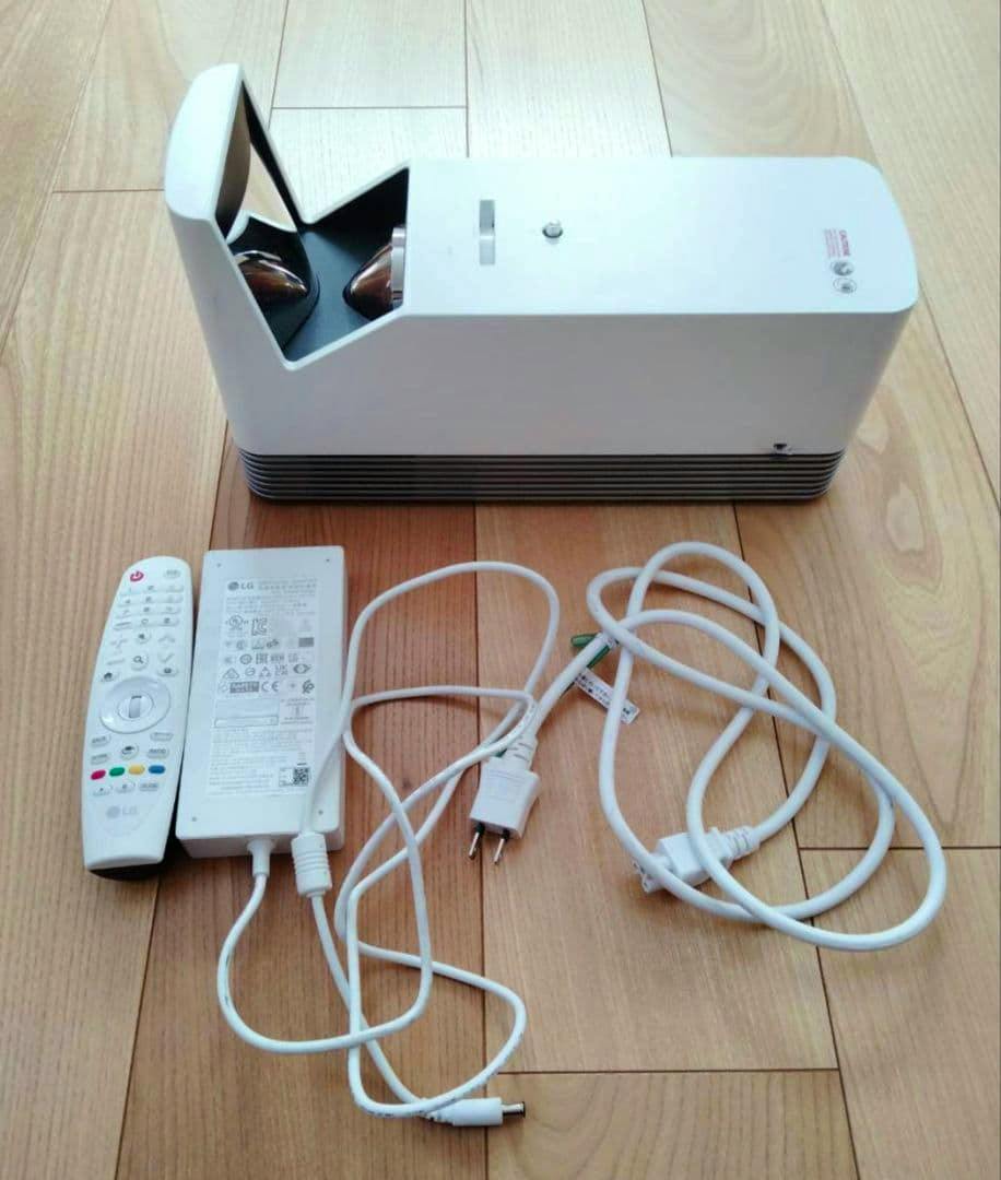 LG HF85LSR projector main unit shown with its remote and power cables laid out on a wooden floor.