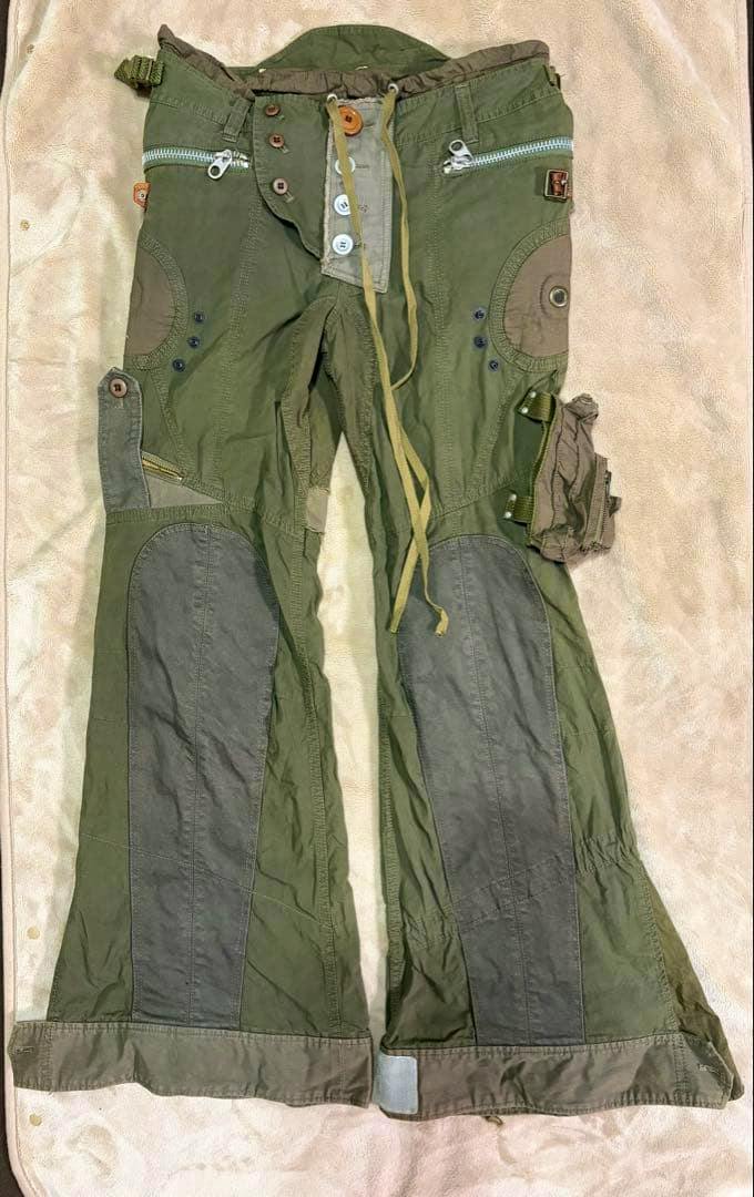 Front view of olive green men's cotton cargo pilot pants laid flat showing zipper pockets, button fly, drawstring waist, and reinforced knee panels.