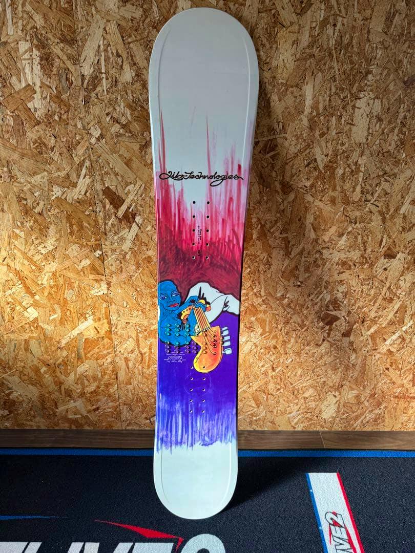 Lib Technologies (Jamie Lynn model) snowboard standing vertically against a plywood wall, showing purple and pink top-sheet graphics and binding mount holes.