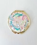 Gold-rimmed compact mirror showing a decorative pastel illustration on the cover.