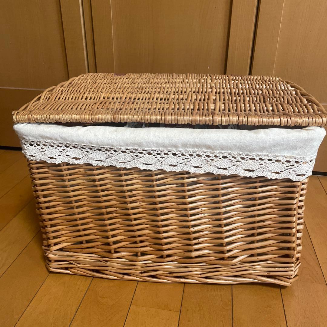 Woven straw storage basket with lid and white lace fabric trim sitting on a wooden floor.