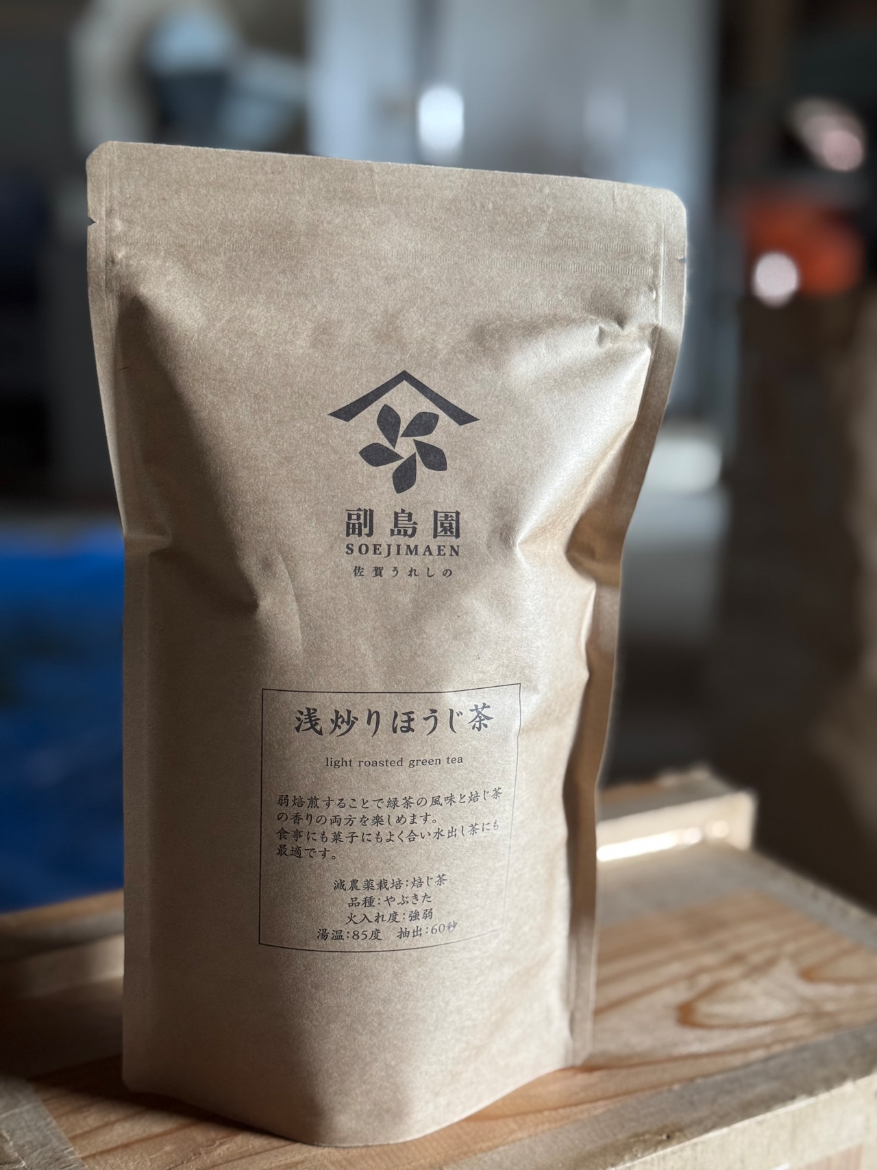 A kraft paper pouch labeled 浅炒りほうじ茶 (loose-leaf green tea) with a printed logo and text, standing on a wooden surface.
