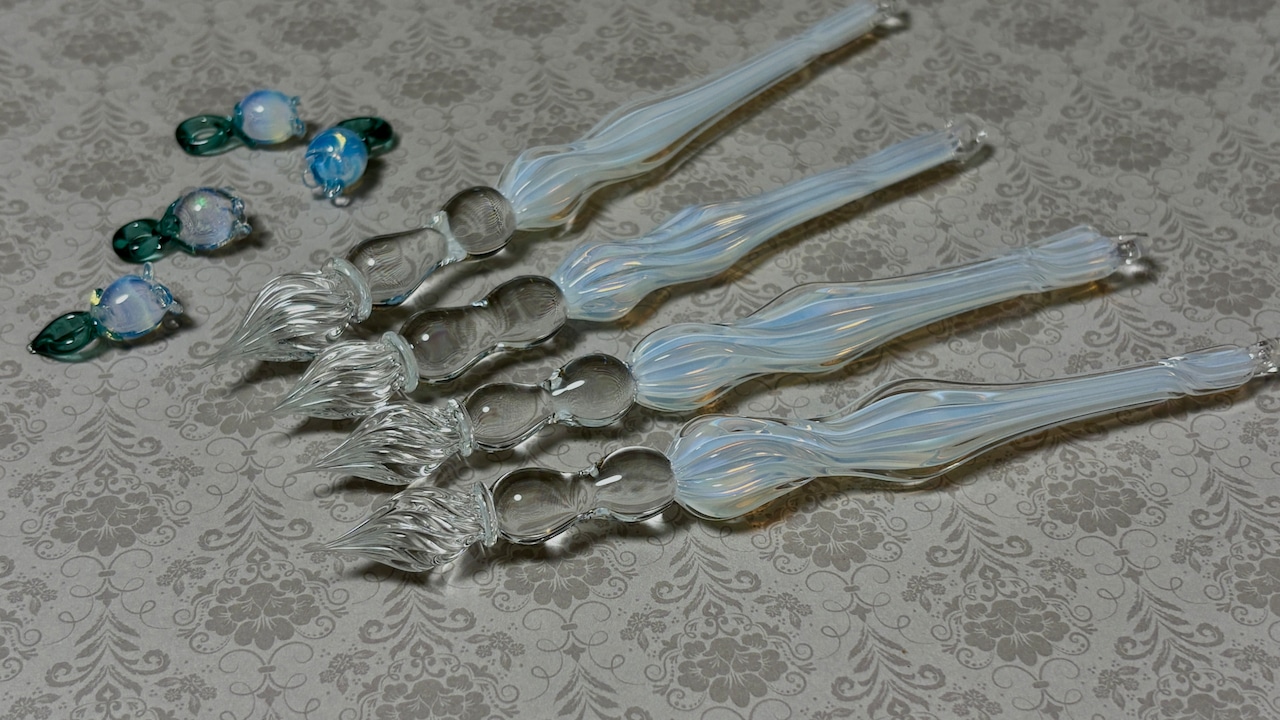 Five glass fountain pens with matching small glass charms arranged on a patterned surface.