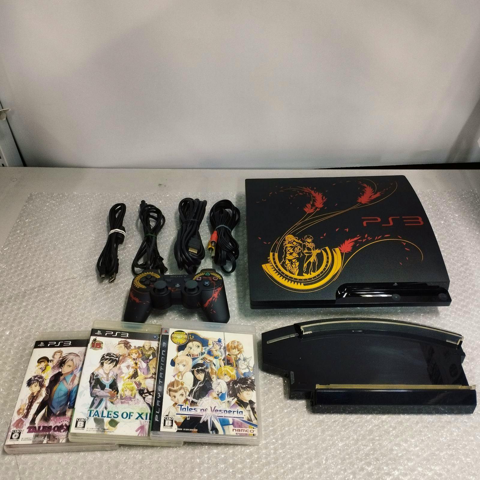 A PS3 console with Tales-themed artwork, a matching controller, cables, and several PS3 game cases on bubble wrap.