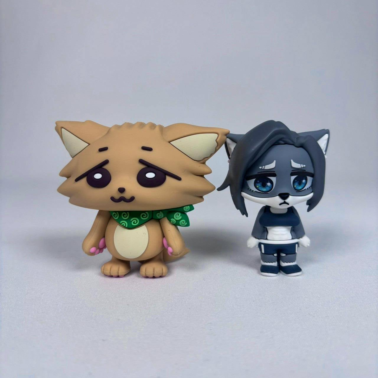 Two sofubi plastic figures standing on a white surface, a tan fox-like figure with a green scarf on the left and a gray wolf-like figure with blue eyes on the right.
