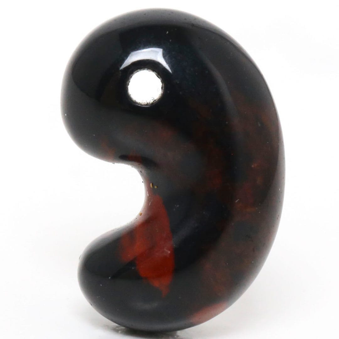 Close-up photo of a comma-shaped magatama pendant stone with a drilled hole, dark with red mottling, on a white background.