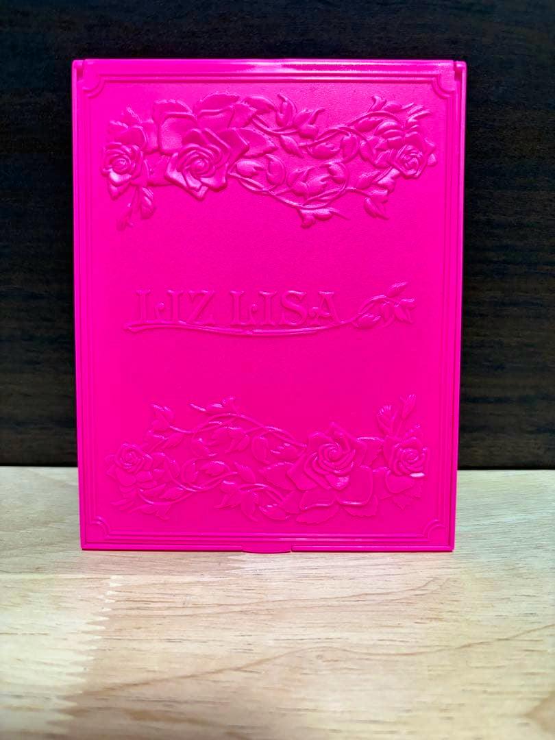 Pink LIZ LISA compact mirror with embossed rose decorations and the brand name on the front, shown standing on a wooden surface.