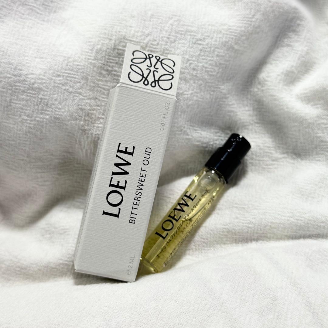 A 2ml LOEWE Bittersweet Oud glass perfume vial with black cap beside its white LOEWE box on a white textured fabric background.
