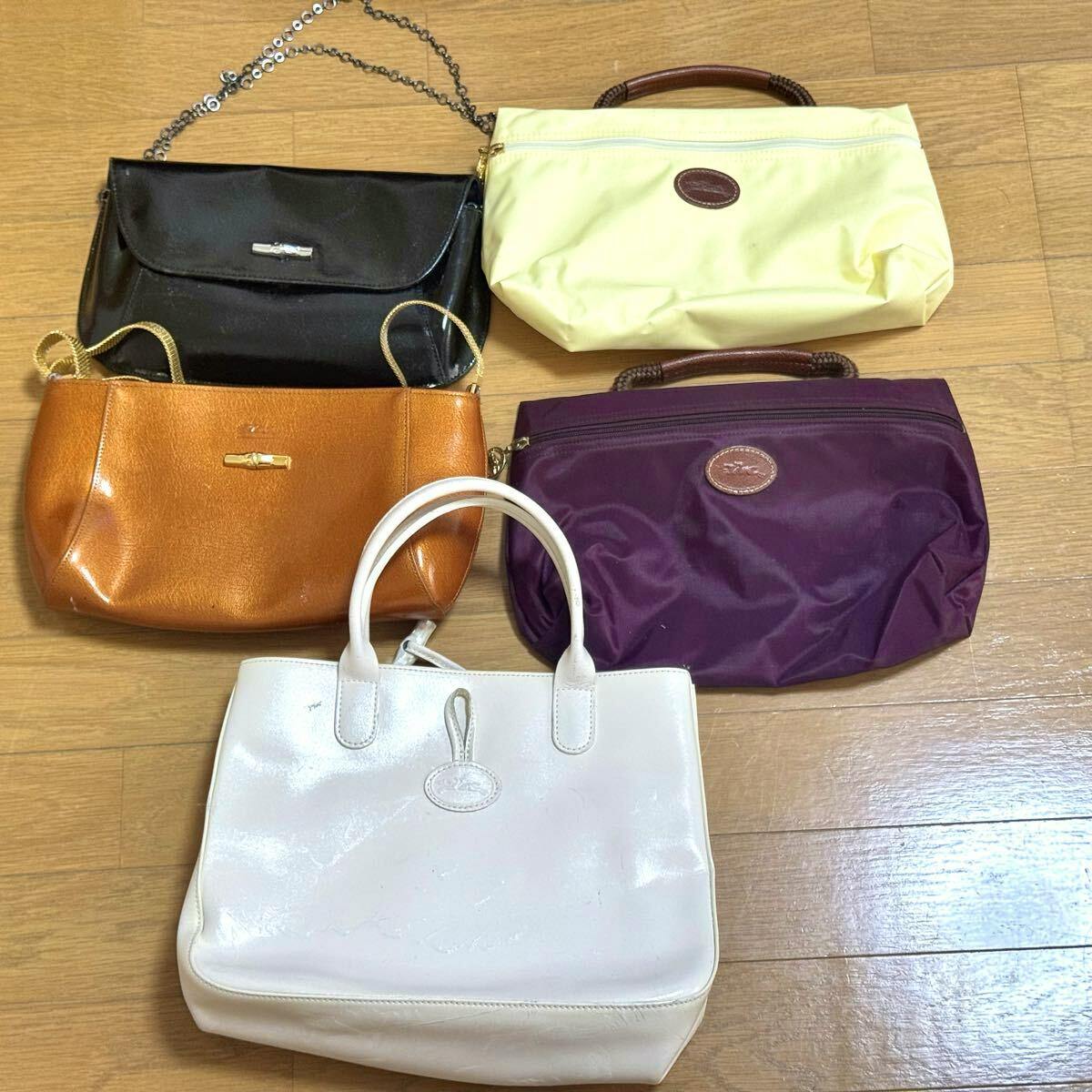 Five Longchamp bags and pouches (white tote, brown shoulder bag, black patent clutch, pale yellow pouch, purple nylon pouch) displayed on a wooden floor.