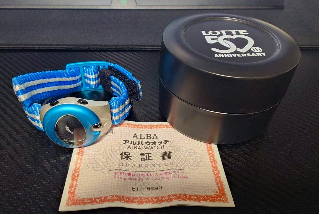 Mint-blue ALBA watch with a blue striped fabric strap pictured next to a round black Lotte 50th Anniversary case and a guarantee/warranty paper.