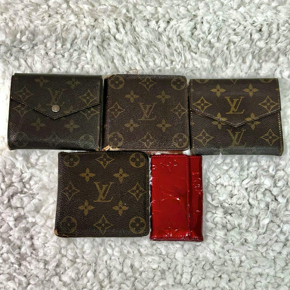 Five small Louis Vuitton monogram wallets and a red patent Vernis piece arranged on a white textured rug.