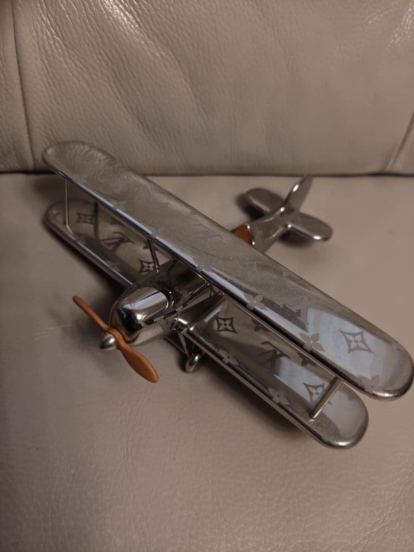 An iron ornamental airplane paperweight with Louis Vuitton monogram pattern and a small wooden propeller shown on a beige leather surface.