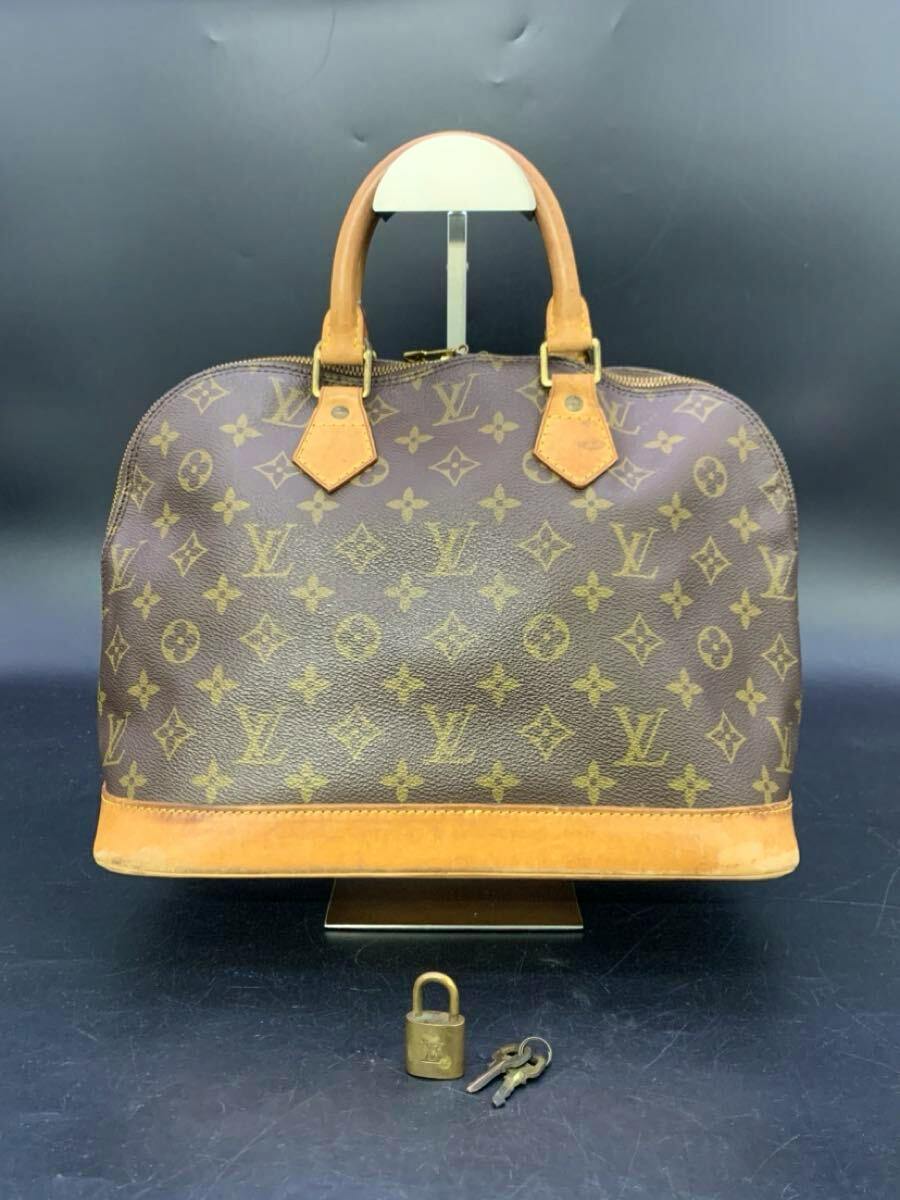 Front view of the Louis Vuitton Alma monogram canvas handbag with leather trim, handles, and a padlock with keys placed in front.
