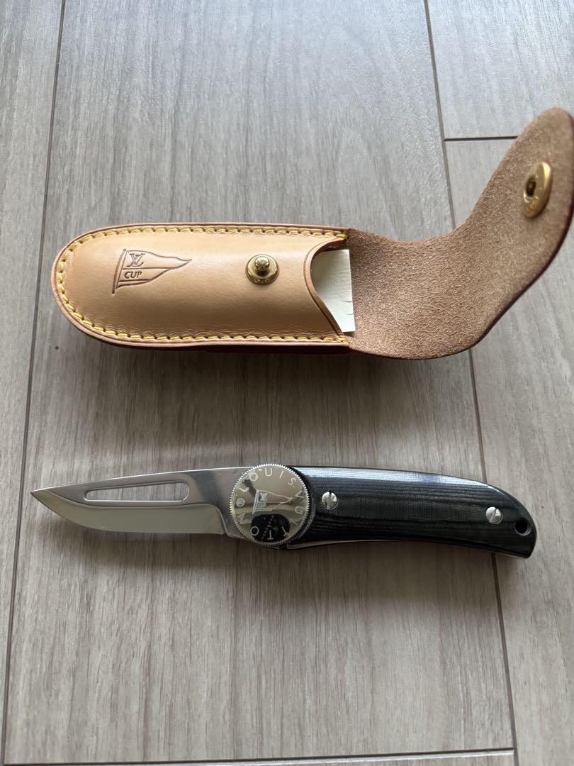 Top-down photo of the Louis Vuitton Americas Cup pocket knife with blade extended beside its tan leather sheath on a wood floor.