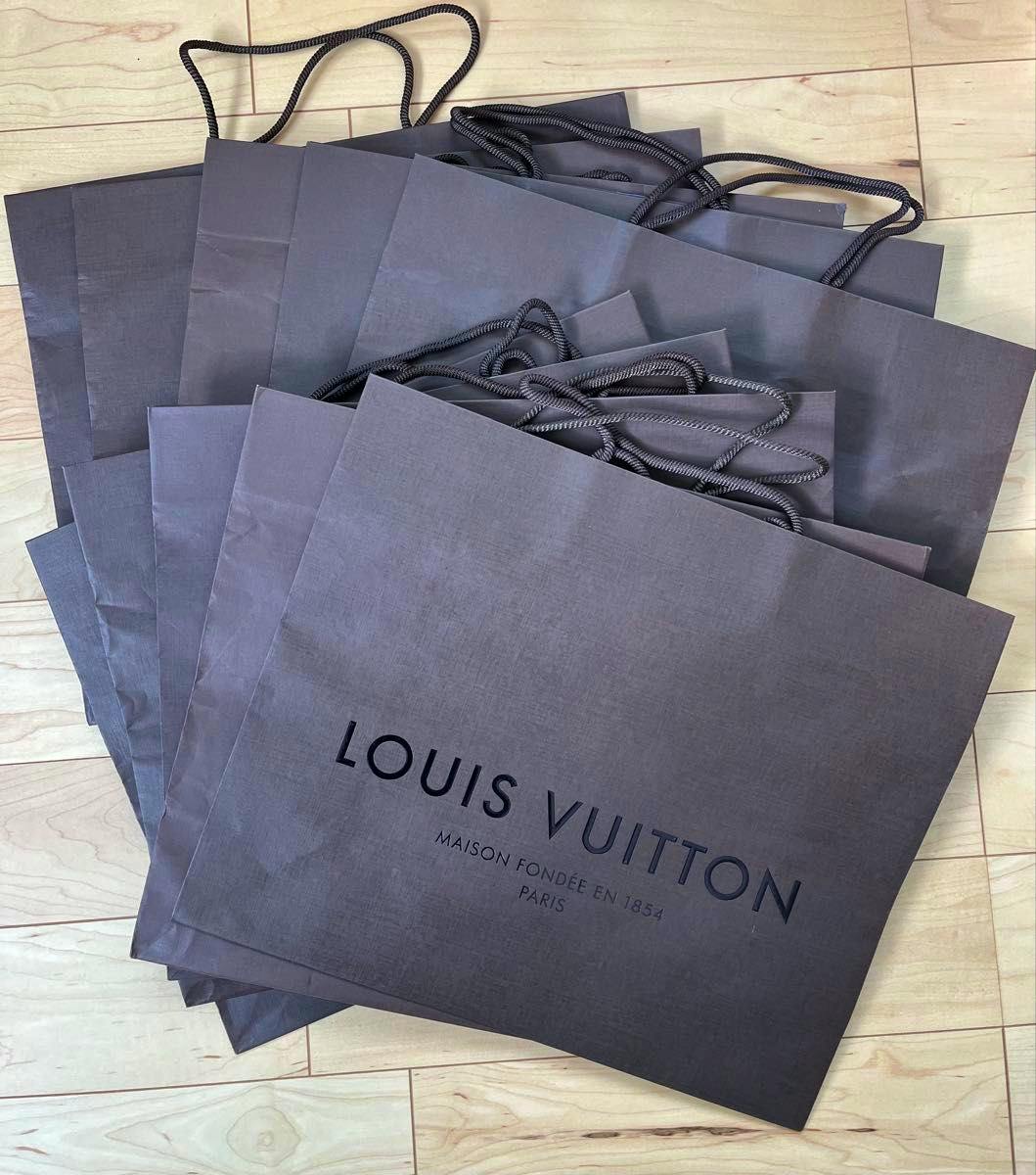 Stack of brown Louis Vuitton paper shopping bags with rope handles arranged on a light wood floor, the front bag showing the LOUIS VUITTON logo.
