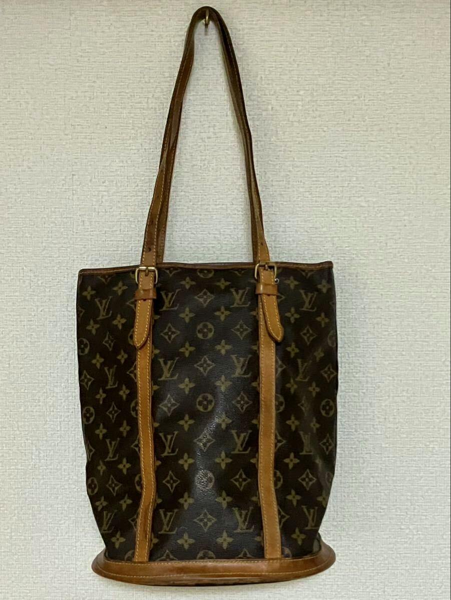 Front view of the Louis Vuitton Bucket GM bag in monogram canvas with tan leather trim and shoulder straps hanging against a wall.