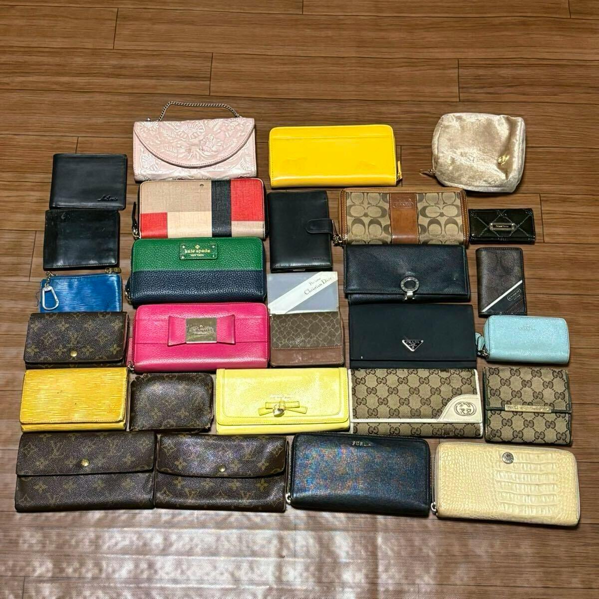 Flat lay of 28 assorted wallets, key cases, and small pouches including Louis Vuitton and other designer brands arranged on a wooden floor.