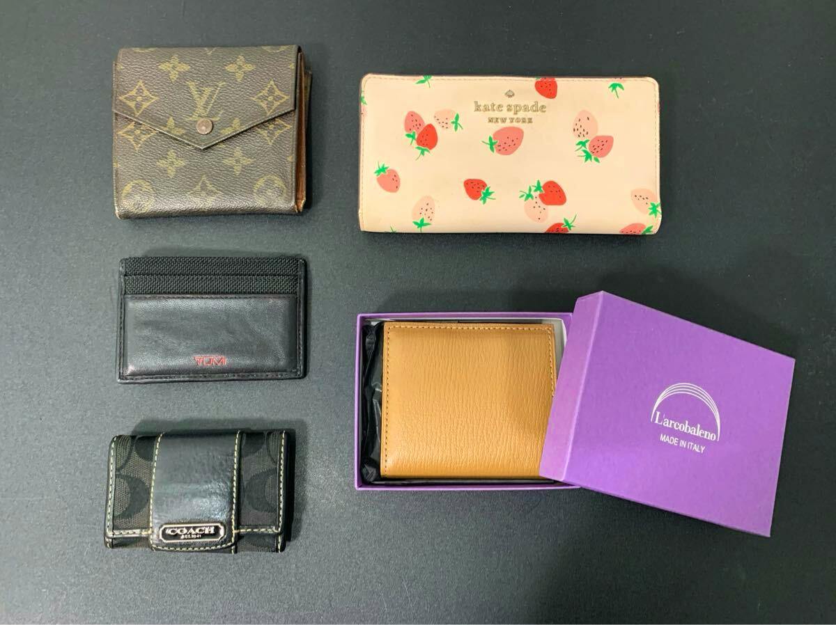 Five wallets and cases (Louis Vuitton monogram, Kate Spade strawberry wallet, TUMI card holder, COACH key case, and a tan L'arcobaleno wallet in a purple box) arranged on a dark surface.