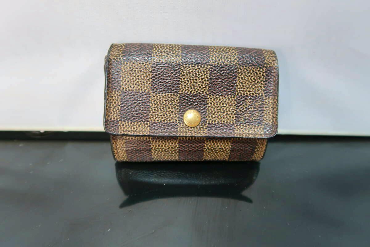 A closed Louis Vuitton Damier canvas coin case shown front-facing with a gold snap button on a black surface.