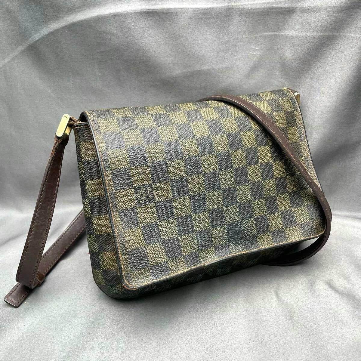 Louis Vuitton Damier Ebene Musette Tango shoulder bag with brown leather strap displayed on a gray background.
