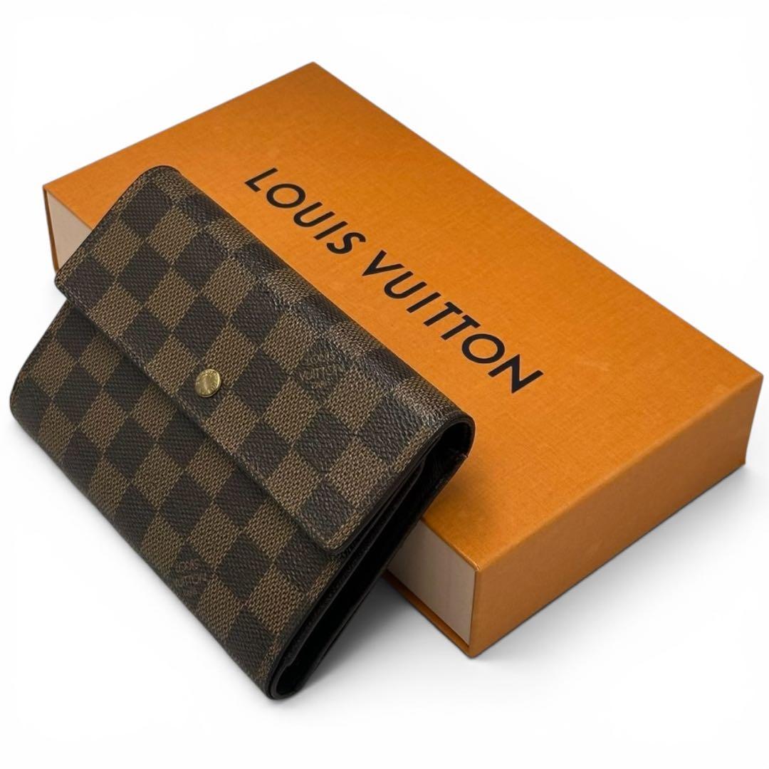 Damier-pattern three-fold Louis Vuitton wallet resting on an orange Louis Vuitton box.