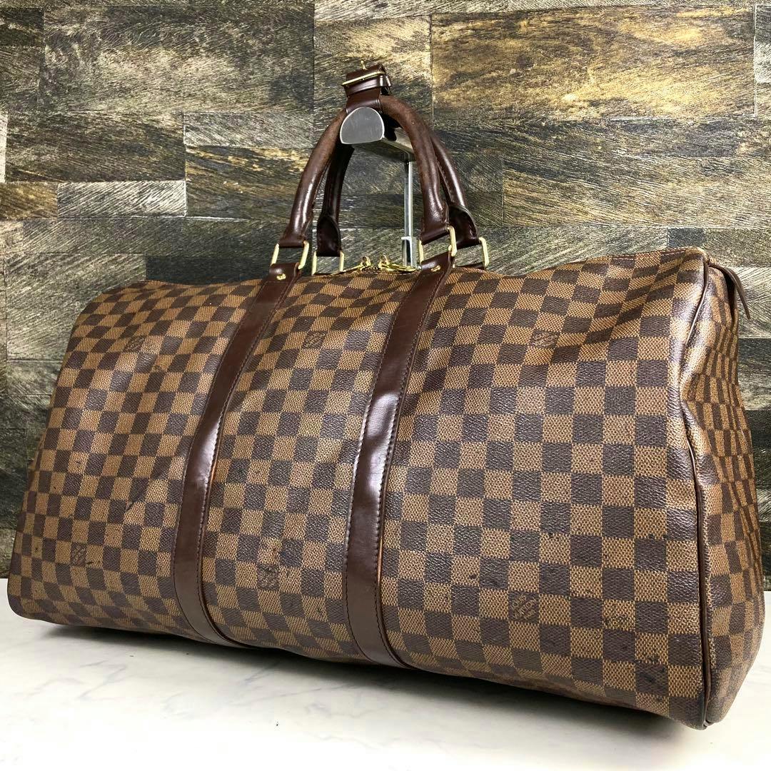 Brown Louis Vuitton Damier Ebene duffle bag with dark brown leather straps and gold hardware shown against a stone background.