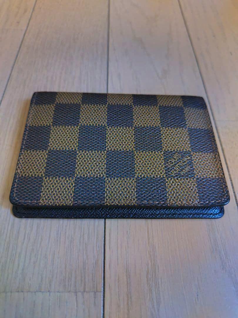 Closed Louis Vuitton Damier card/pass case on a wooden floor showing the front checkered canvas and edge.