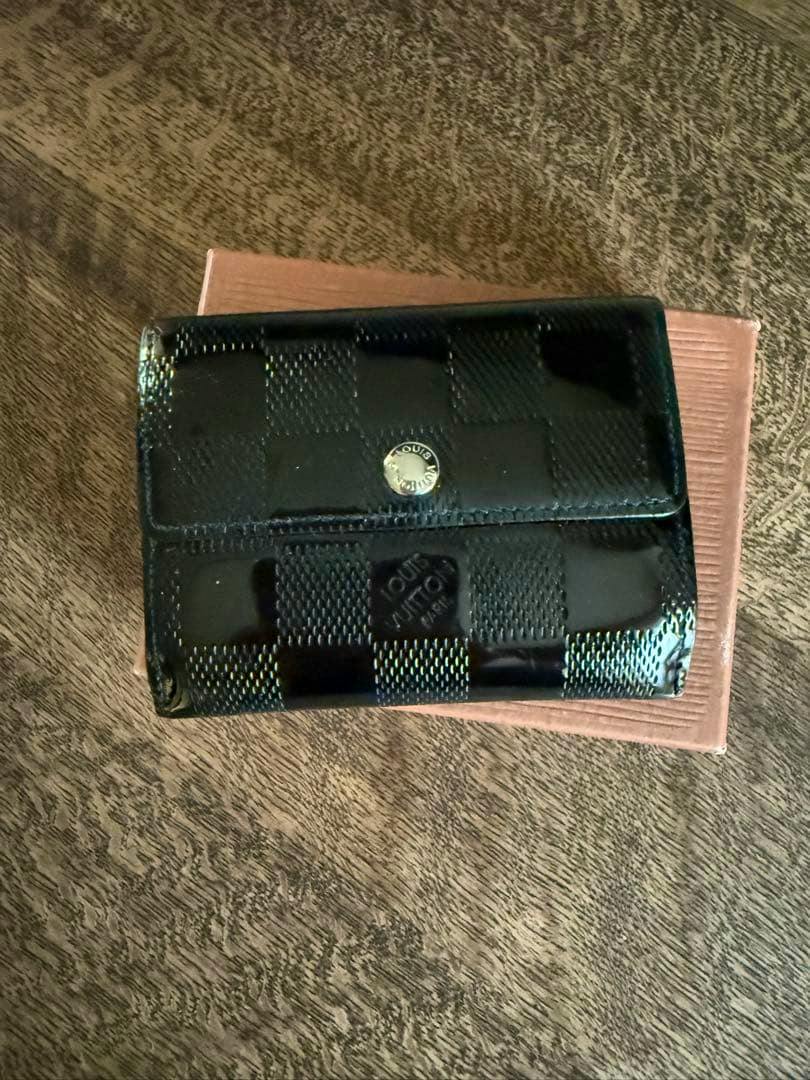 Closed black Louis Vuitton Damier Vernis Ladoux coin case (patent leather, M92136) placed on a box.