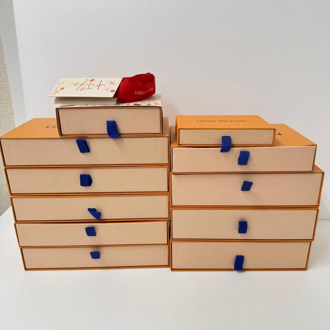 Stacked Louis Vuitton orange drawer boxes with blue ribbon pulls and a small red pouch and tag on top.