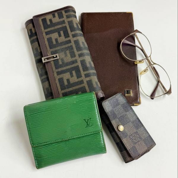 Five branded accessories arranged on a white background: a green Louis Vuitton wallet, a Fendi long wallet, a Damier key case, a brown glasses case, and a pair of eyeglasses.