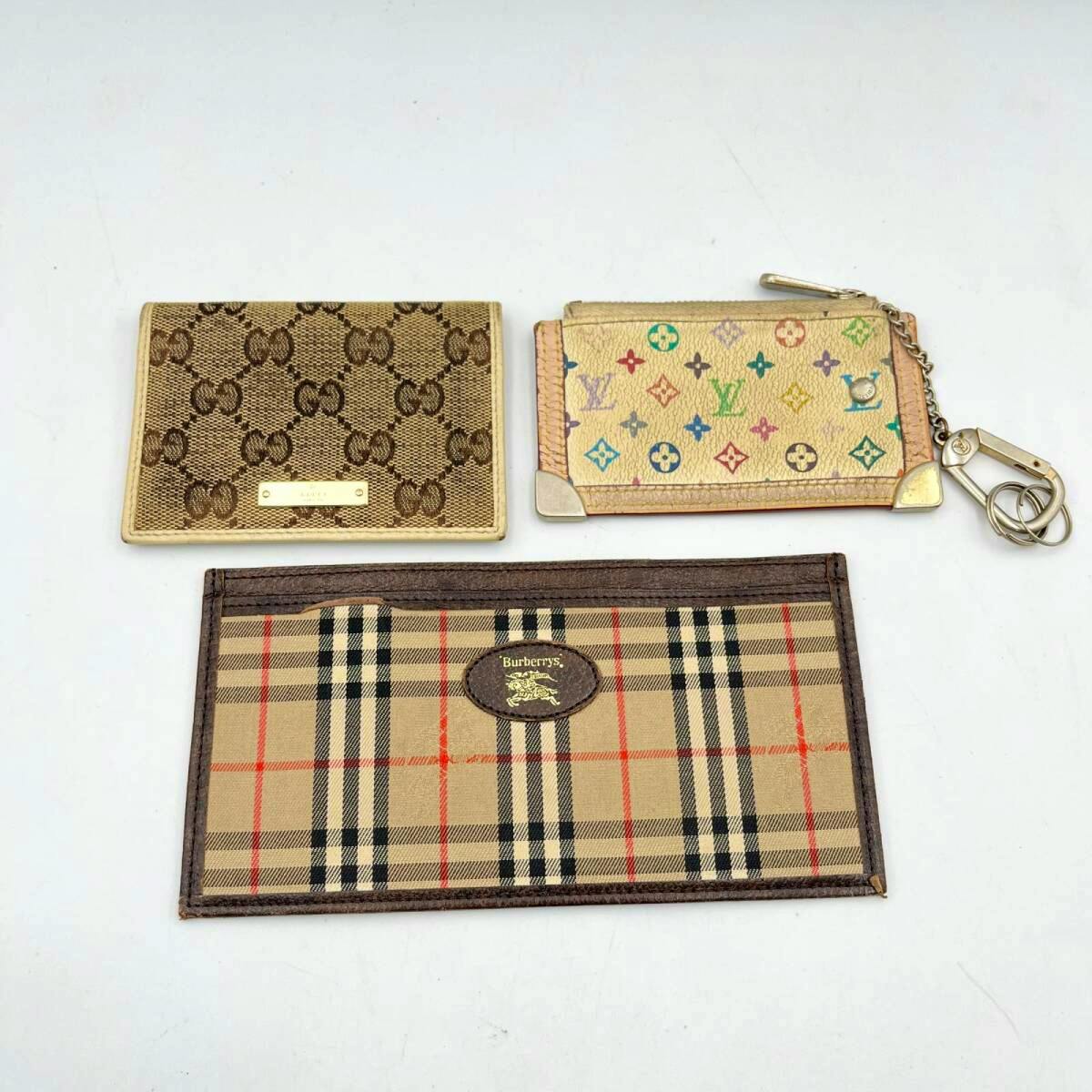 Three small canvas wallets/card cases on a white background: a Gucci monogram card case, a Louis Vuitton multicolor coin/key pouch with chain, and a Burberry check card holder.