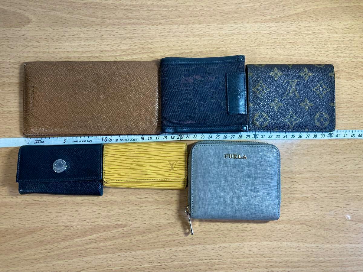 Top-down photo of several wallets and small leather goods, including a Louis Vuitton monogram piece and a Furla zip wallet, arranged on a wooden table with a measuring tape.