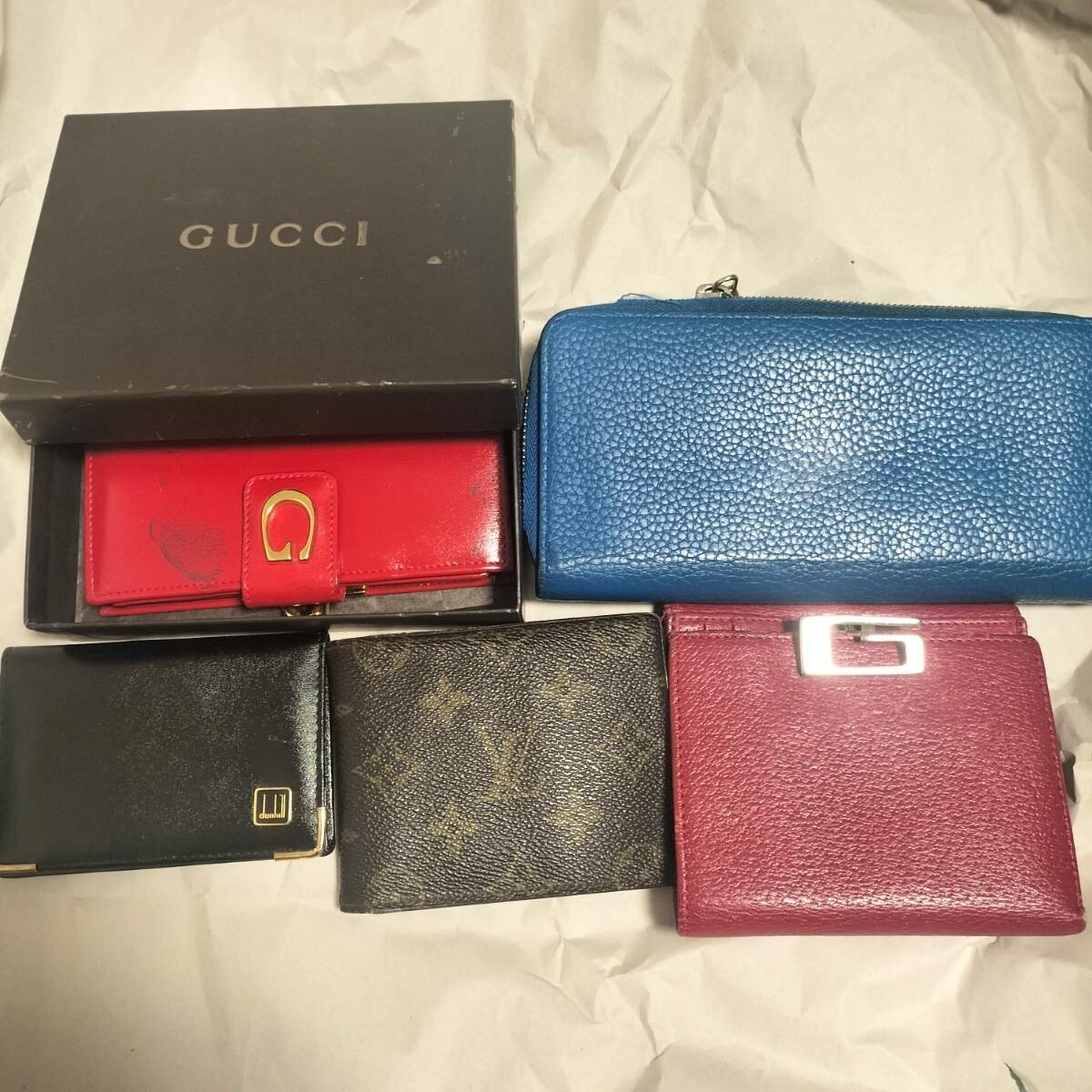 A collection of wallets and a Gucci box on a surface, including a blue zip-around wallet, a red wallet in a box, a brown monogram wallet, and two other wallets.