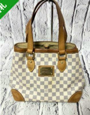 Front view of the Louis Vuitton Hampstead shoulder bag in checkered canvas with brown leather trim and a rectangular metal nameplate.