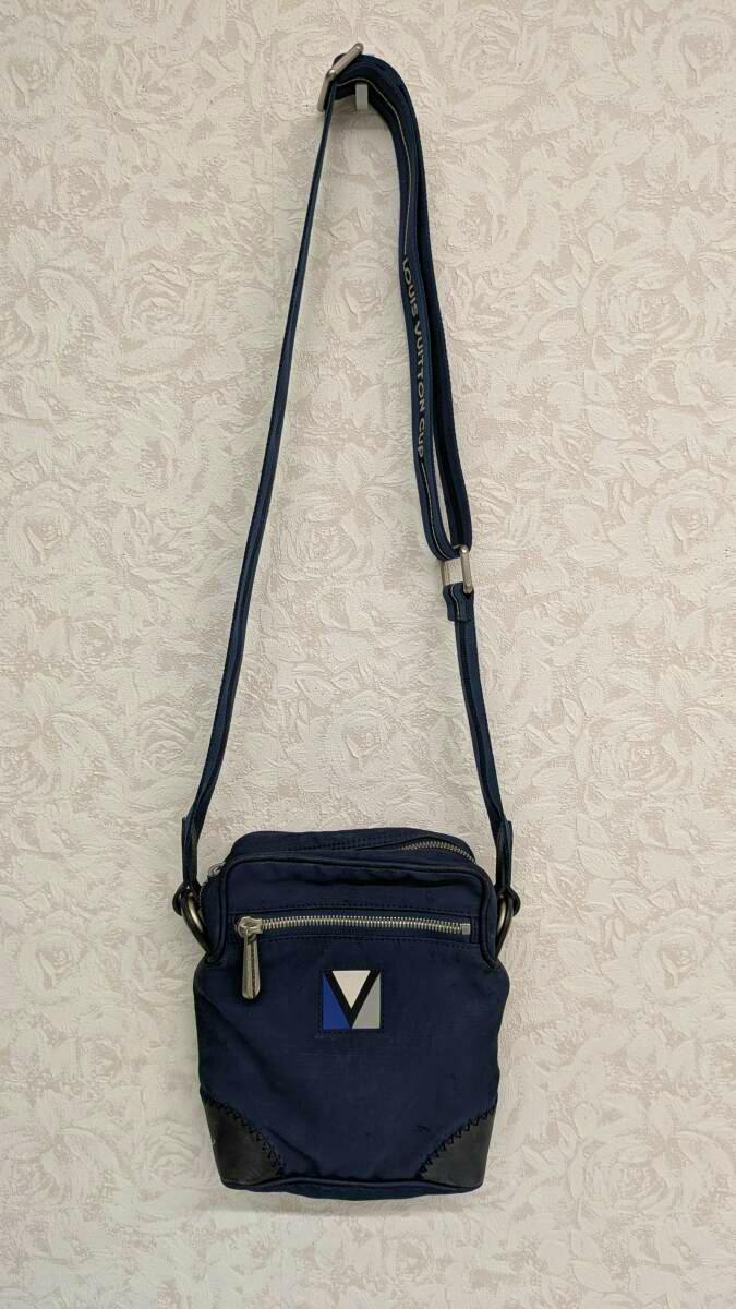 Front view of a navy Louis Vuitton canvas shoulder bag with a front zip pocket, LV logo detail, and an adjustable shoulder strap hanging on a wall.