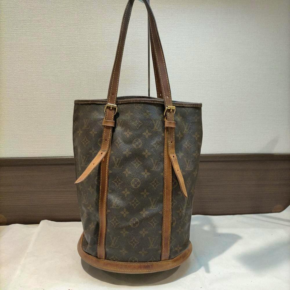Front view of a brown Louis Vuitton monogram bucket bag with leather trim and shoulder straps, shown standing upright.