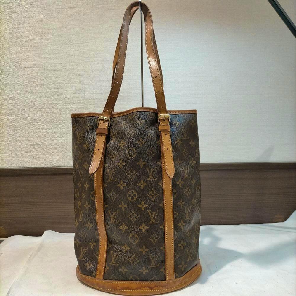 Brown Louis Vuitton monogram handbag with leather trim and two long shoulder straps standing against a neutral background.