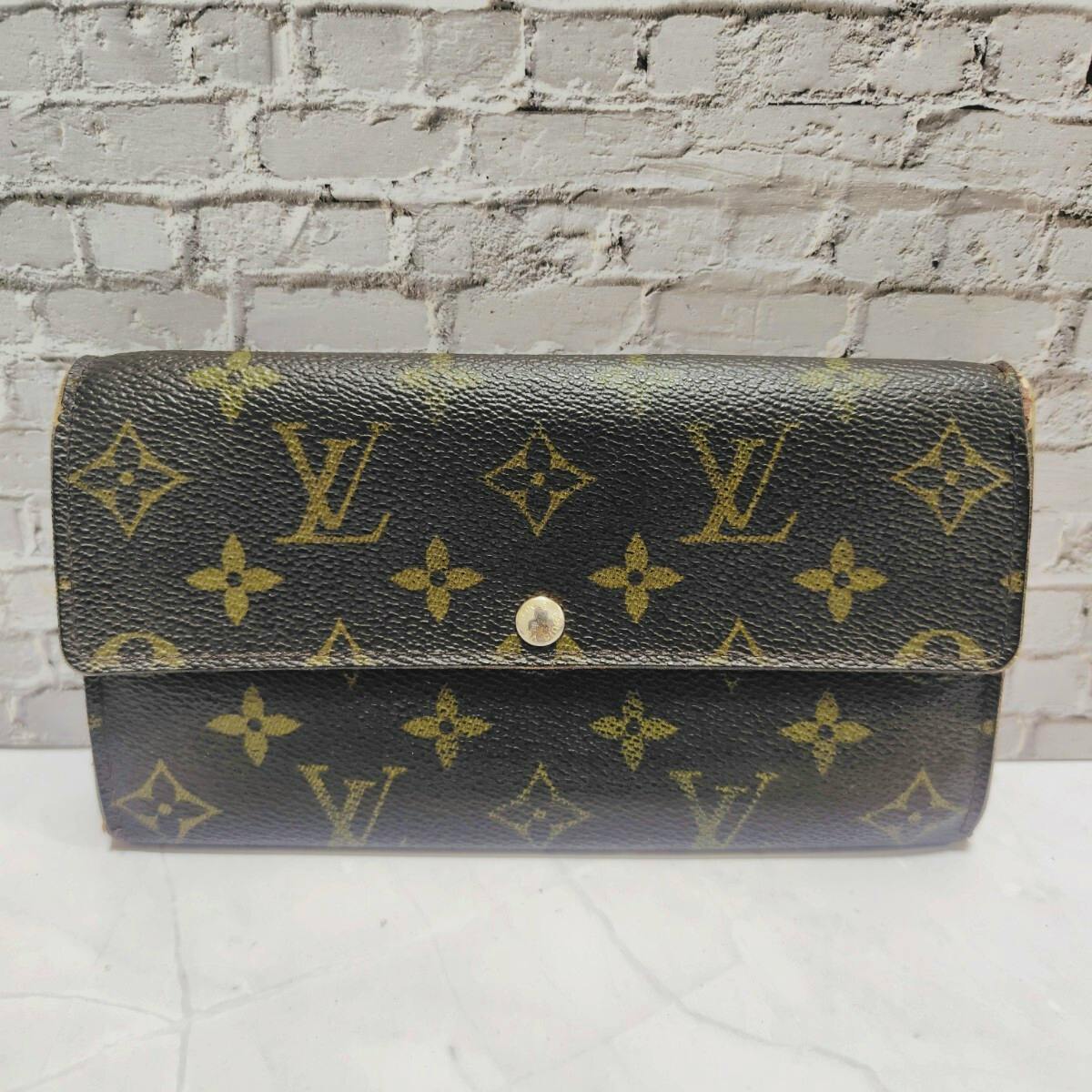 Front view of a Louis Vuitton brown monogram canvas long wallet with a snap button closure on a light brick background.